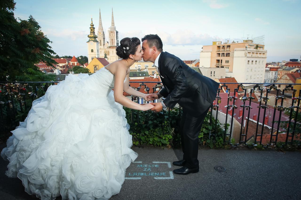 Newlyweds holding hands and kissing in the kissing spot with a view of the cathedral and buildings of Zagreb city in the background.