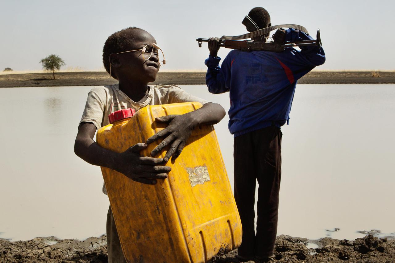 A young boy carries water in a jerry can to his village while his grandfather, the village chief, cradles an AK 47 rifle watching out for cattle rustlers and lions, at the only water source in the area, along the Sobat River in the Greater Upper Nile regi