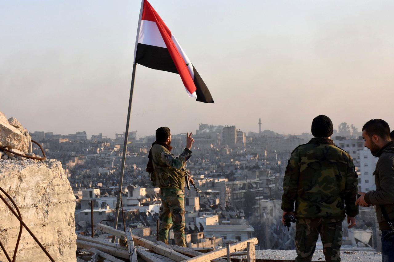 A Syrian government soldier gestures a v-sign under the Syrian national flag near a general view of eastern Aleppo after they took control of al-Sakhour neigbourhood in Aleppo, Syria in this handout picture provided by SANA on November 28, 2016. SANA/Hand
