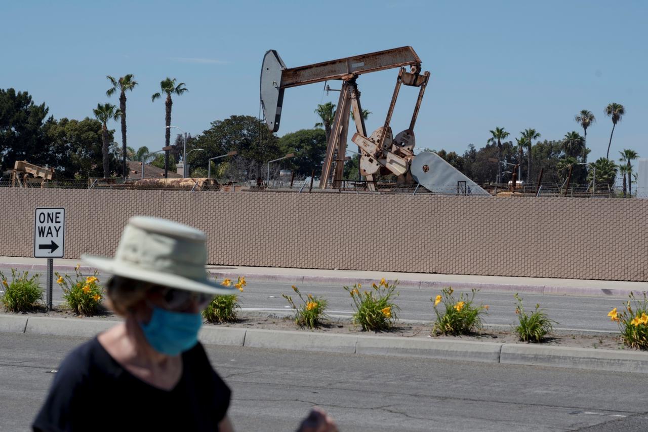 A pedestrian wearing a protective mask walks past an oil derrick in Huntington Beach