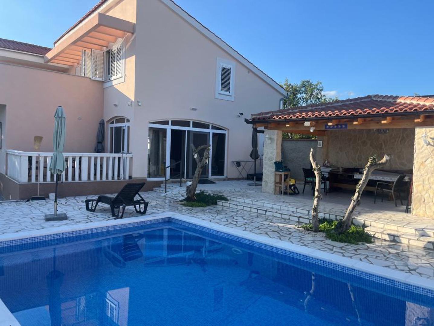 Vanjski prostor kuće zamišljen je za uživanje i opuštanje. Na raspolaganju je uređeni vrt, pergola s nadstrešnicom za sjedenje, prostor za roštilj, sunčalište i veliki betonski bazen obložen keramikom, opremljen suvremenom tehnikom za održavanje kvalitete vode, toplinskom pumpom i solarnim pokrovom. 