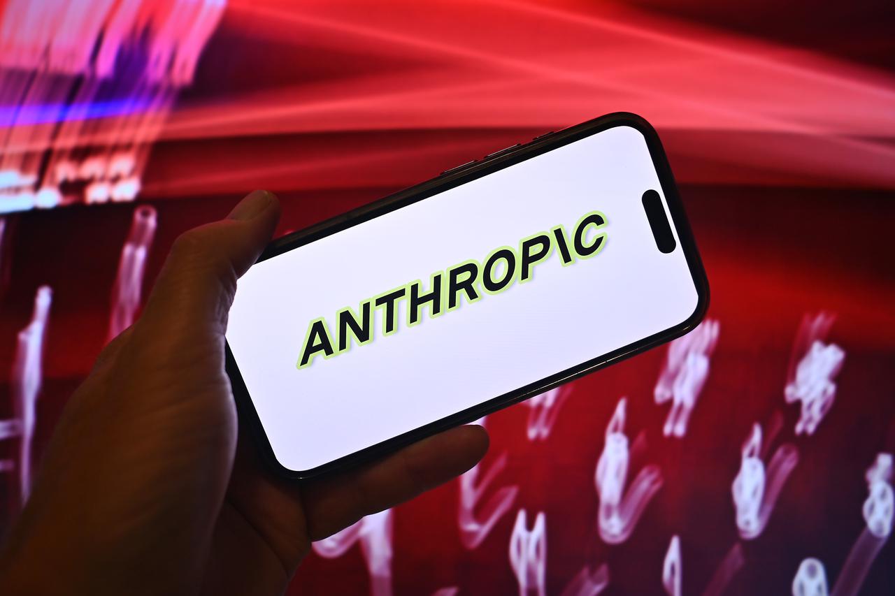 Hand shows a smartphone with the Anthropic logo in front of a computer background