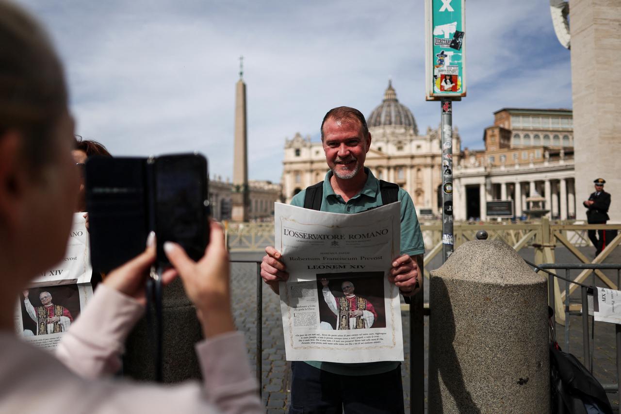 A woman takes a photo of a man holding Vatican newspaper L'Osservatore Romano containing a photograph of the newly elected Pope Leo XIV, at the Vatican