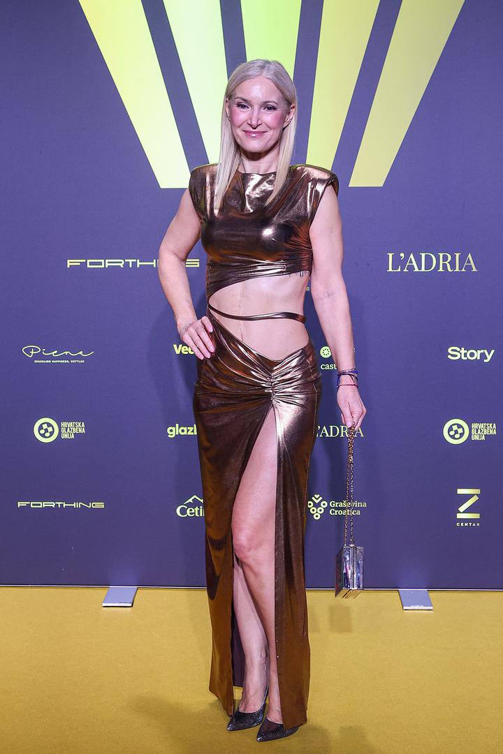 Uršula je odjenula zlatnu asimetričnu haljinu visokog sjaja, a haljina je otkrila njezin seksi trbuh te duge i vitke noge. Odabrala je profinjenu šminku te je kosu raspustila.