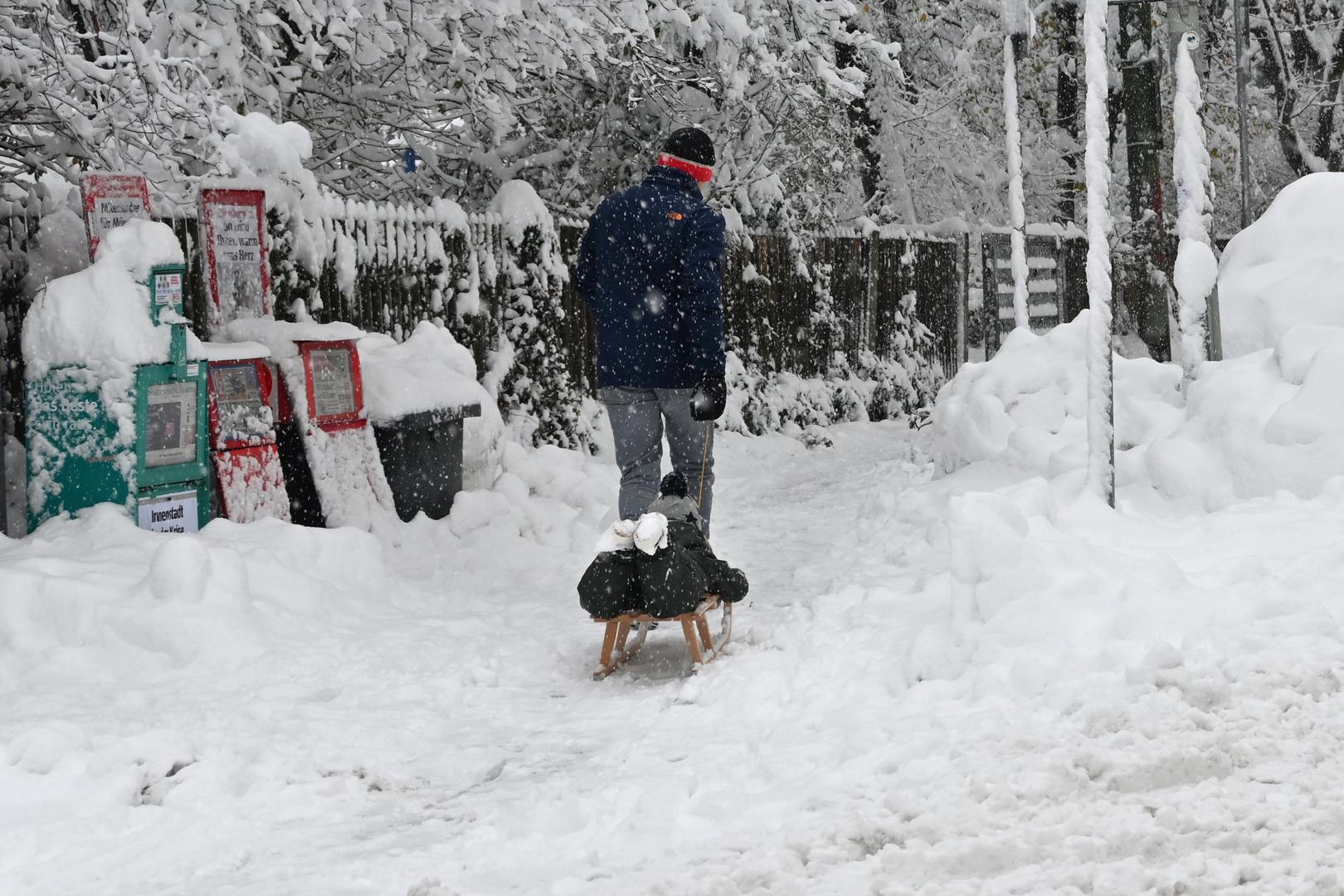 02 December 2023, Bavaria, Munich: A man pulls a child on a sledge along a snow-covered sidewalk. Snow and ice have caused chaos on the roads and on the railroads in southern Bavaria. Photo: Katrin Requadt/dpa Photo: Katrin Requadt/DPA